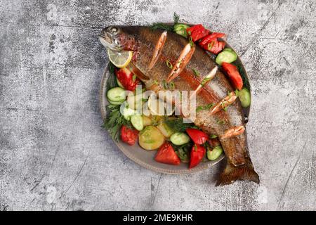 Im Ofen gebackene Regenbogenforelle mit Gemüse und Gemüse auf einem runden Teller auf dunklem Hintergrund. Draufsicht, flach liegend Stockfoto
