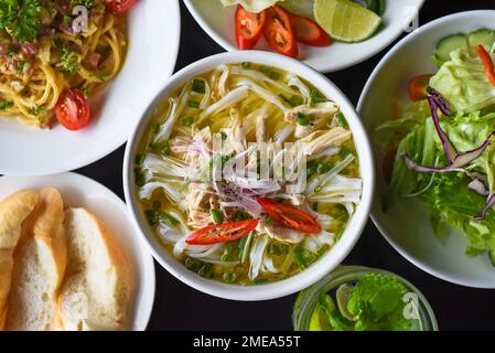 Hühnersuppe Pho ga im vietnamesischen Stil auf schwarzem Hintergrund mit Banh mi Brot, Salat und Pasta Carbonara Stockfoto