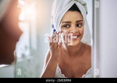 Auf dem Weg zur strahlenden Haut. Eine attraktive junge Frau, die sich im Bad Feuchtigkeitscreme ins Gesicht trägt. Stockfoto