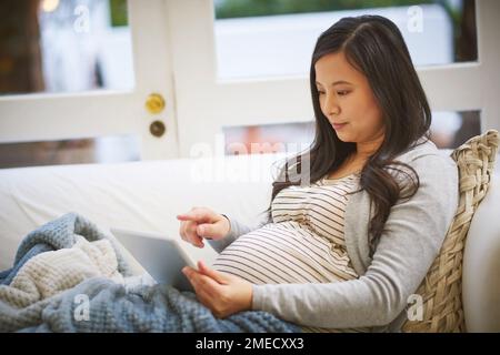 Ich recherchiere so viel wie möglich für den großen Tag. Eine schwangere Frau, die zu Hause ein digitales Tablet benutzt. Stockfoto