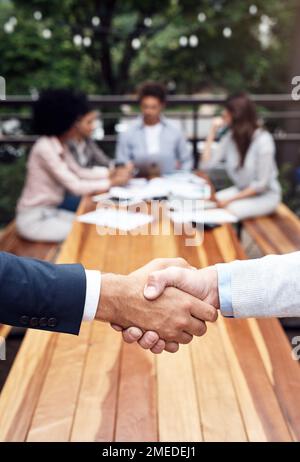 Es war mir ein Vergnügen, mit Ihnen Geschäfte zu machen. Zwei unbekannte Geschäftsmänner, die sich die Hand schütteln, während ihre Kollegen hinter ihnen draußen sitzen. Stockfoto