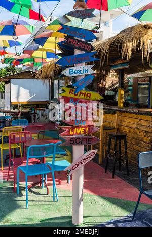 Ksamil, Albanien - 11. September 2021: Straßenschild aus Holz auf dem Café mit Wegbeschreibungen zu verschiedenen Orten der Welt unter bunten Schirmen i. Stockfoto