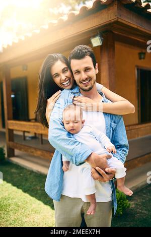 Und drei ergibt eine Familie. Abgeschnittenes Porträt einer glücklichen dreiköpfigen Familie draußen mit ihrem Haus im Hintergrund. Stockfoto