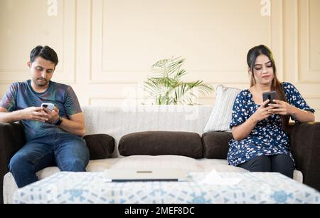 Ein junges indisches Paar, das zu Hause mit dem Handy beschäftigt ist, während es in der Ferne auf dem Sofa sitzt - Konzept der Technik-, Smartphone- und Social-Media-sucht. Stockfoto