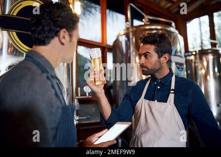 Das ist unsere beste Ladung bisher. Zwei fokussierte junge männliche Unternehmer testen tagsüber eine frische Charge Bier in ihrer Bierbrauerei. Stockfoto