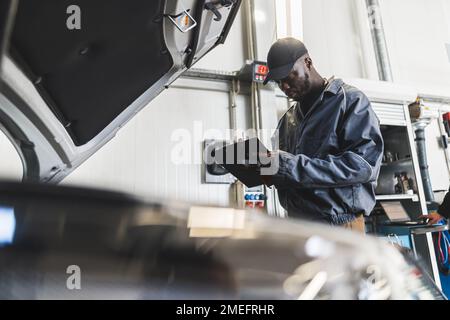 Ein schwarzer Mechaniker, der Sachen auf seinem Block überprüft. Verschwommener Vordergrund. Werkstattkonzept. Hochwertiges Foto Stockfoto