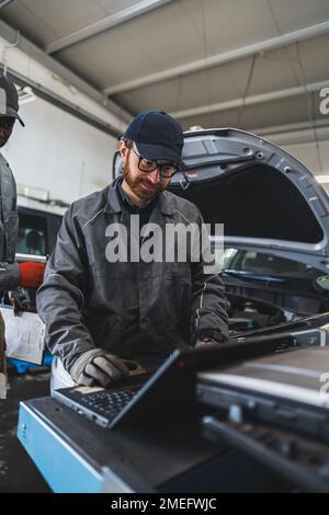 Vertikales Foto. Mittlere Aufnahme. Ein weißer, männlicher Mechaniker, der eine Computerdiagnose in einer Werkstatt durchführt, und ein anderer Mechaniker steht hinter ihm. Hochwertiges Foto Stockfoto