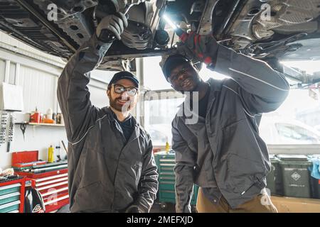 Mittellange Aufnahme von zwei Mechanikern, die während der Reparatur des Fahrgestells an die Kamera lächeln. Werkstattkonzept. Hochwertiges Foto Stockfoto