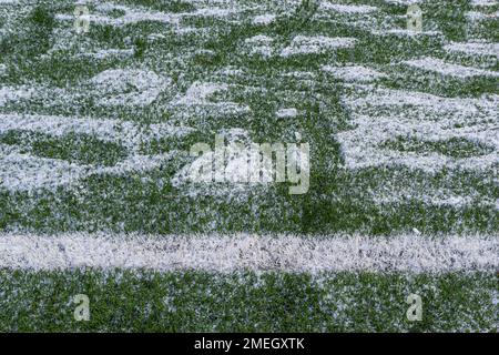Das Fußballfeld mit Kunstrasen ist mit einer leichten Schneeschicht bedeckt. Frühling. Grünes Gras auf dem Fußballfeld ist von zu sehen Stockfoto