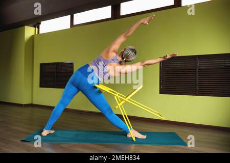Eine Frau führt einen gesunden Lebensstil und praktiziert Yoga, führt eine Variation der Trikonasana Übung mit Unterstützung auf einem Stuhl, Dreieck Pose, trai Stockfoto