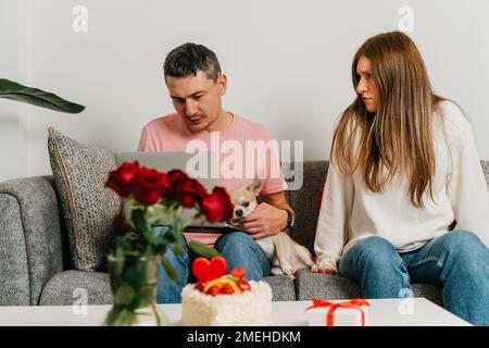 Wunderschönes junges Paar mit Hund, das auf der Couch sitzt und sich über ein romantisches Date am Valentinstag streitet. Ein Mann, der am Laptop arbeitet, eine Frau im Zorn Stockfoto