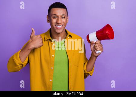 Foto eines netten jungen Mannes zeigt Daumen hoch halten Megafon Ankündigung gekleidet stilvolles gelbes Kleidungsstück isoliert auf violettem Hintergrund Stockfoto