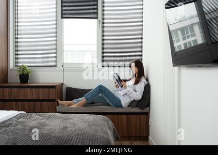 Frau liest Buch in einem Schlafzimmer mit Bett und Holzgarderobe in einem modernen Apartment Stockfoto