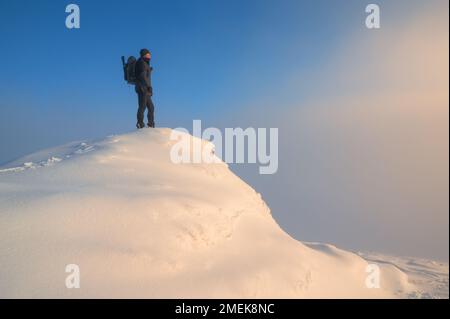 Einsamer Wanderer auf dem Berggipfel, der an einem schneebedeckten Wintertag einen Wurm bei Sonnenuntergang sieht Stockfoto