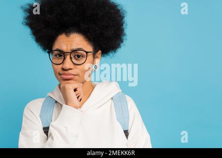 Verärgerte und rücksichtsvolle afroamerikanische Schülerin mit Brille, die Hand am Kinn hält und isoliert auf Blau auf die Kamera schaut Stockfoto