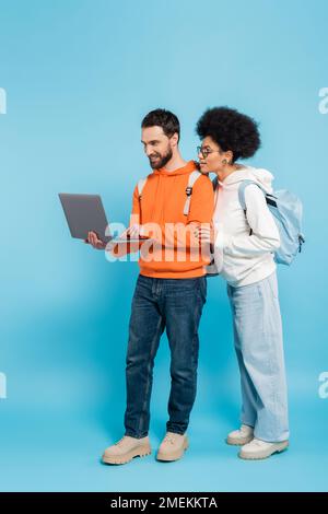 Volle Länge eines bärtigen Schülers, der einen Laptop in der Nähe einer afroamerikanischen Freundin auf Blau benutzt Stockfoto