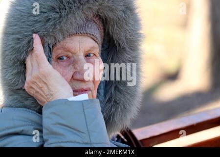 Porträt einer älteren Frau im warmen Mantel mit Pelzhaube, die auf einer Holzbank im Park sitzt. Alter, kaltes Wetter, Leben im Ruhestand Stockfoto