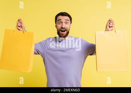 Ein bärtiger Mann in einem lila Pullover, der Einkaufstaschen in der Hand hält und isoliert auf Gelb auf die Kamera schaut Stockfoto