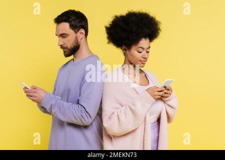 Trendige Kommunikation zwischen verschiedenen ethnischen Paaren auf Mobiltelefonen, während Sie auf Gelb isoliert von Rücken zu Rücken stehen Stockfoto