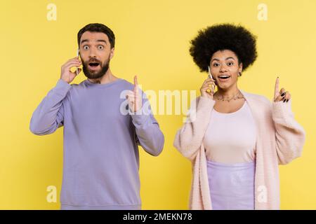 Erstaunliches, multiethnisches Paar, das eine Geste mit Ideen zeigt, während es sich auf einem gelben, isolierten Smartphone unterhält Stockfoto