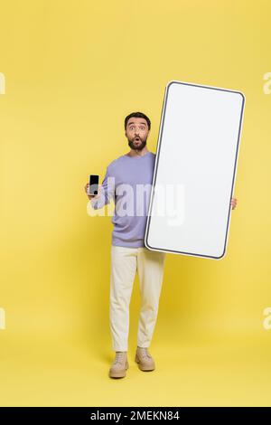 Die ganze Länge eines überraschten Mannes, der ein Smartphone mit einem leeren Bildschirm in der Nähe einer weißen Vorlage für ein Mobiltelefon auf grauem Hintergrund zeigt Stockfoto