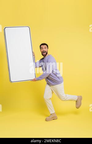 Ein bärtiger Mann in voller Länge, der in die Kamera schaut, während er mit einer Handyvorlage auf grauem Hintergrund läuft Stockfoto