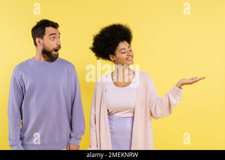 Lächelnde afroamerikanische Frau, die mit der Hand auf einen bärtigen Mann zeigt, isoliert auf Gelb Stockfoto