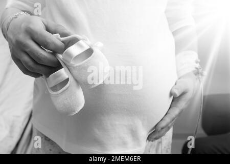 Kleine Schuhe für das ungeborene Kind im Bauch der Schwangeren. Schwangere Frau hält Schuhe für kleines Baby Entspannen im Krankenhaus vor der Geburt, glückliche Schwangerschaft Stockfoto