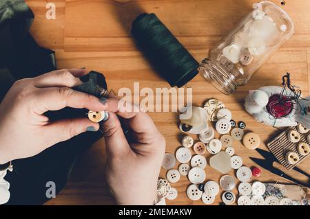 Ansicht von oben mit weiblichen Händen, die einen Knopf auf ein schwarzes Tuch aufsticken, mit Nähgeräten auf einem Holztisch und Kopierbereich. Stockfoto