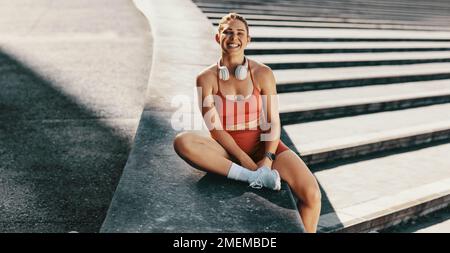 Sportlerin sitzt an der Wand und lächelt in die Kamera. Sportliche Frau, die eine Pause vom Laufen im Freien macht. Glückliche Frau, die morgens trainiert. Stockfoto
