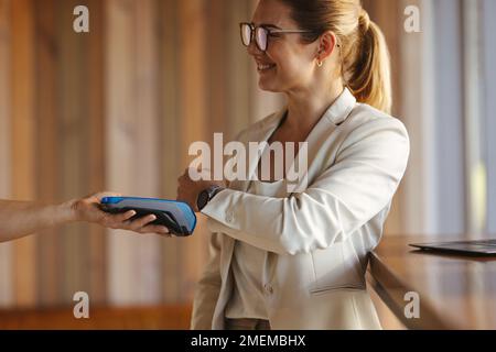 Eine professionelle Frau bezahlt eine Rechnung mit einer Smartwatch in einem Café. Glückliche Geschäftsfrau, die eine kontaktlose Zahlung mit NFC-Technologie tätigt. Stockfoto