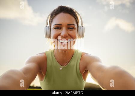 Fröhliche Sportlerin, die draußen ein Selfie macht. Die Frau, die mit Kopfhörern die Kamera anlächelt, ist fit. Eine Frau, die sich beim morgendlichen Workout amüsiert. Stockfoto