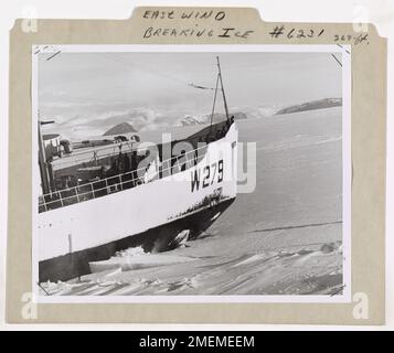 USCGC Eastwind (WAGB-279) Antarktis, Operation Deep Freeze. Eine Nahaufnahme zeigt, wie der verstärkte Bug der 6.515 Tonnen schweren US Coast Guard Eisbrecherin Eastwind auf eine dicke Eishütte im kristallgepflasterten McMurdo Sound fährt und das Eis durch den Sturz ihres Gewichts brechen lässt. Zwei Heckpropeller liefern 10.000 PS bei voller Geschwindigkeit für den Aufstieg. Das Eis kann 7 bis 15 Fuß oder tiefer sein. Manchmal führt die Oberflächenreibung dazu, dass der Bug schnell am Eis klebt. So dass sich der Eisbrecher lösen und wieder vom Sims abheben kann. In diesem Fall wird ihr System von Ferging-Tanks in Bewegung gesetzt. Tausende von Stockfoto