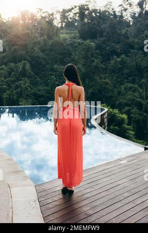 Rückansicht einer jungen Frau, die am Außenpool des Luxusresorts steht. Weibliches Modell im Sonnenkleid. Stockfoto