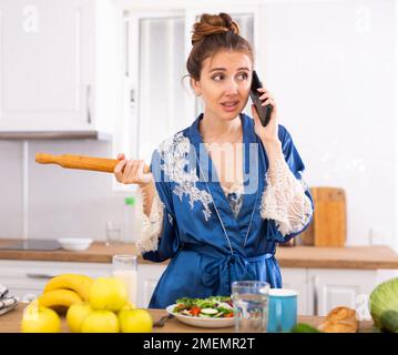 Unzufriedene Hausfrau mit einem Nudelholz in den Händen, die in der Küche auf dem Mobiltelefon sprach Stockfoto