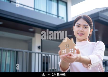 Junge asiatische Frau in Holzform mit Dorfhintergrund, Neuhaus, Hausversicherung, Immobilien oder Wohnungsbaukreditkonzept. Stockfoto