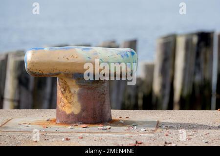 Alte Anlegestelle an einem verlassenen Pier. Das Festmachen ist der Typ, um den eine Seilschlaufe gelegt wird. Er hat alte Farbe und ist rostig. Überreste eines alten Docks dahinter. Stockfoto