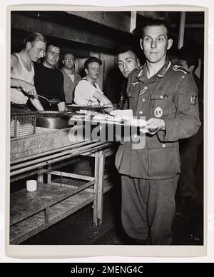 Ein deutscher Gefangener an Bord eines von der Küstenwache bemannten Transportschiffs sieht erstaunt aus, als er zusieht, wie ein Mitgefangener ein beladenes Tablett mit Lebensmitteln erhält. In der Normandie gefangen genommen, werden diese Soldaten, die wahrscheinlich monatelang ohne ordentliche Mahlzeiten unterwegs waren, zur Internierung in die Vereinigten Staaten gebracht. Das Bild spiegelt die Bedingungen wider, denen Kriegsgefangene gegenüberstanden, und die Rolle der Küstenwache in der Kriegslogistik. Stockfoto