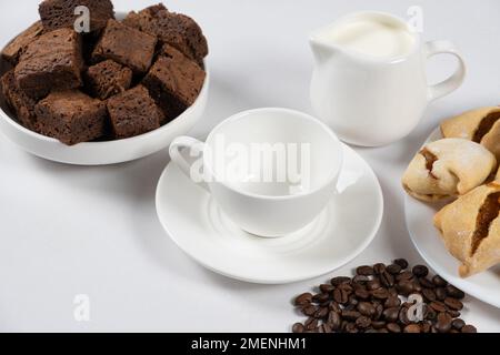 Leere Kaffeekeramilietasse mit Milchkanne, Schokoladenkuchen und Kaffeebohnen auf weißem Hintergrund Stockfoto