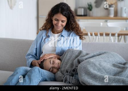Ein wunderbares, bezauberndes Mädchen, das in eine Decke gewickelt ist, liegt auf den Kniekehlen der Frau auf der bequemen Heimcouch Stockfoto