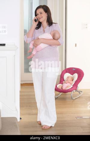 Mutter hält ein Zwillingsbaby (3 Monate) in Armen, während sie am Telefon spricht und das andere Baby im Hintergrund im Wippe schläft Stockfoto