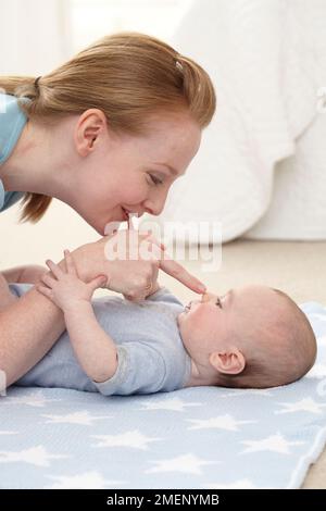 Mutter berührt die Nase eines kleinen Jungen (16 Wochen), der auf dem Boden auf seinem Rücken liegt, Seitenansicht Stockfoto