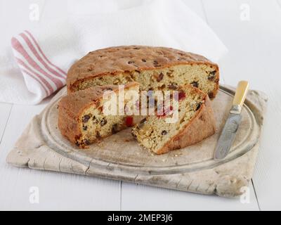 Kirsch- und Rosinenobstkuchen auf Holzbrett mit Scheiben, Messer und Geschirrtuch auf dem Tisch Stockfoto