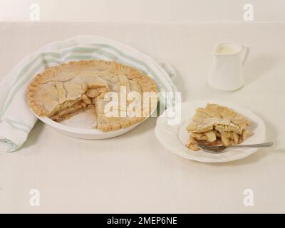Apfelkuchen mit Scheiben auf dem Teller mit Gabel und Kanne mit Sahne im Hintergrund Stockfoto