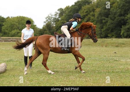 Ein Mädchen reitet Kastanienpony weg vom Baumstamm, nachdem es während des Reitstages in der Koppel darüber gesprungen ist, während die Lehrerin zusieht, mit Blick auf die Seite Stockfoto