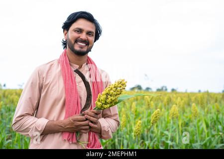 Glücklicher, lächelnder junger Landwirt, der die Ernte und Sichel hält, indem er vor die Kamera auf das Mais- oder Maisfeld schaut - Konzept von echten Landbewohnern, Glück und Anbau Stockfoto