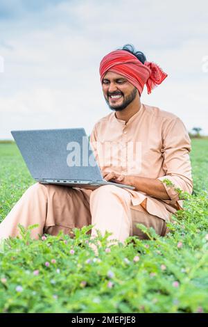 Vertikale Aufnahme eines fröhlichen, lächelnden jungen Landwirts, der auf dem Ackerland mit einem Laptop arbeitet – Konzept von Technologie, moderner Landwirtschaft und Entwicklung Stockfoto