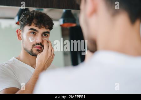 Porträt eines gutaussehenden jungen Mannes, der sein Spiegelbild betrachtet, sein Gesicht berührt, sein Gesicht mit Feuchtigkeitscreme befeuchtet. Über die Schulter. Gesichtspflege Stockfoto
