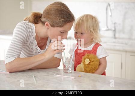 Frau und Mädchen trinken Wasser aus demselben Glas mit Strohhalmen, 18 Monate Stockfoto