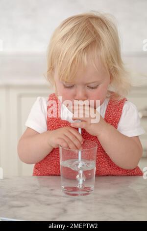 Ein Mädchen trinkt Wasser aus Glas mit einem Strohhalm, 18 Monate Stockfoto